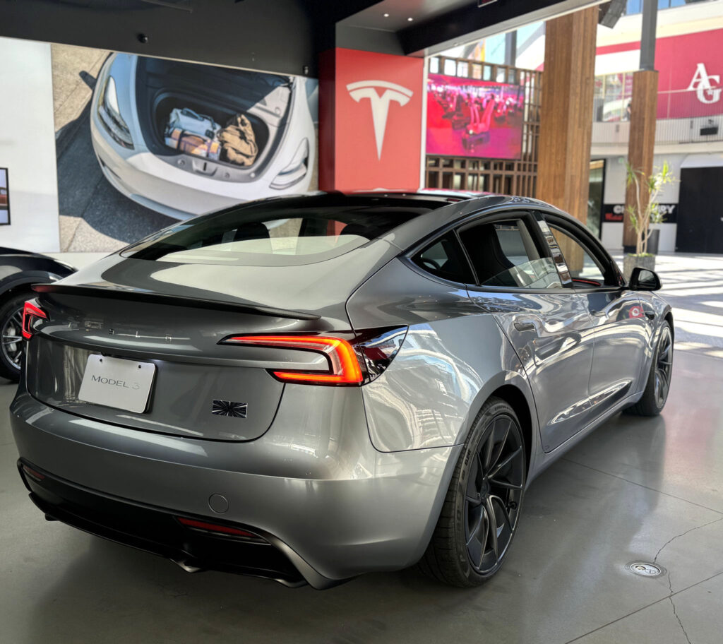 Rear side view of a Tesla Model 3 Highland in Quicksilver color inside a Tesla Store in the United States.